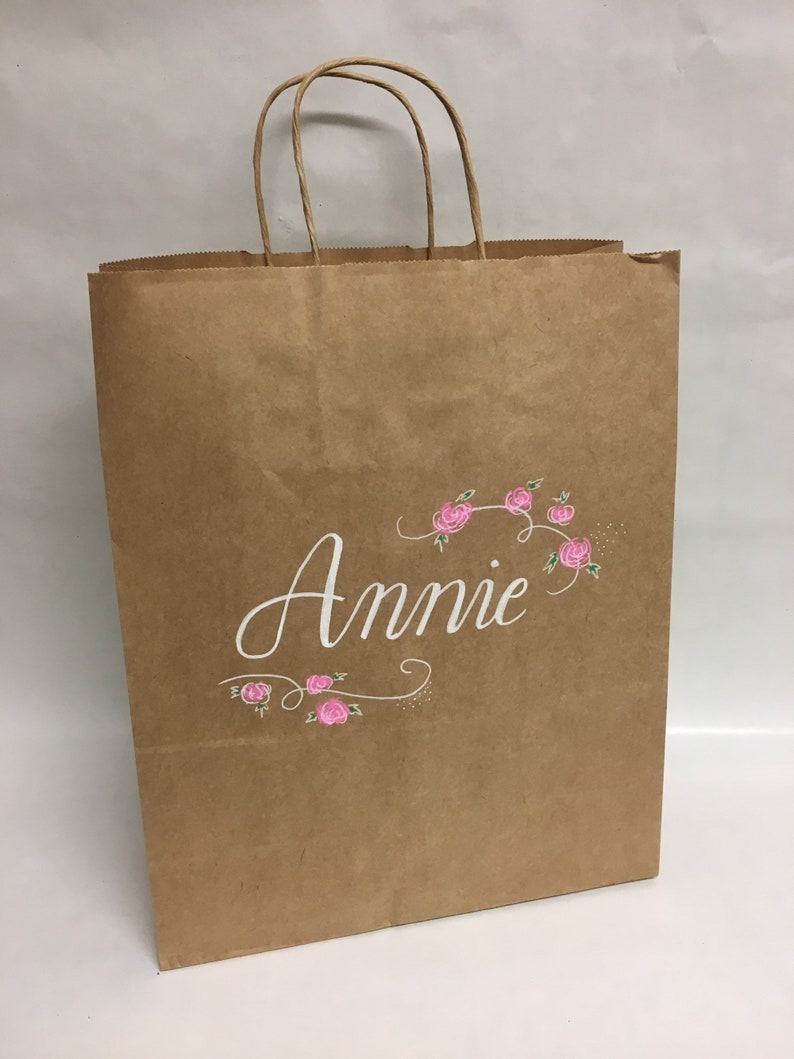 May include: A brown paper shopping bag with twisted paper handles. The word "Annie" is written in white script, with pink floral accents. The bag is designed for gift giving or retail use.