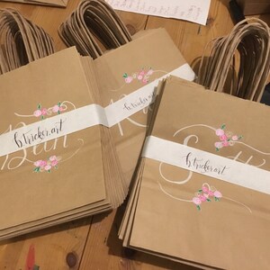 May include: Kraft paper gift bags with white bands featuring cursive text and floral accents. The bags have brown twisted paper handles. The text on the bags reads "b.tricker.art" and "Run" or "S". Pink and white floral designs are painted on the bags.