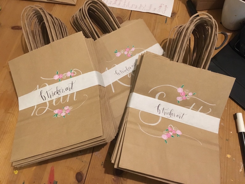 May include: Kraft paper gift bags with white bands featuring cursive text and floral accents. The bags have brown twisted paper handles. The text on the bags reads "b.tricker.art" and "Run" or "S". Pink and white floral designs are painted on the bags.