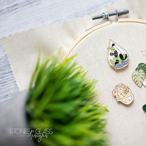 May include: A set of seven enamel pins featuring different potted plants, including a cactus, a succulent, a terrarium, and a bee. The pins are displayed on a white fabric background.