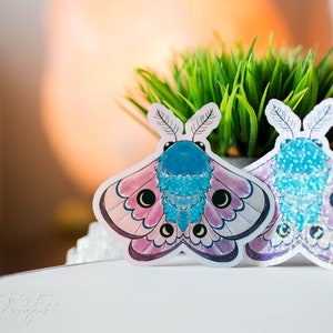 Puede incluir: Dos pegatinas holográficas iridiscentes de una polilla azul y rosa con ojos negros. Las pegatinas están sobre una superficie blanca.