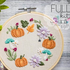 May include: An embroidery hoop with an autumnal design, including orange pumpkins, purple flowers, and green leaves. The words "FULL kit" are visible. A cluster of purple crystals and green foliage are in the background.