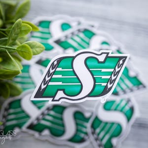 Peut inclure: Plusieurs autocollants avec un logo vert et blanc représentant la lettre "S" et un motif stylisé. Le logo est bordé de noir. Les autocollants sont sur une surface claire, avec un feuillage vert en arrière-plan.