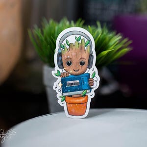 以下が含まれることがあります： ガーディアンズ・オブ・ギャラクシーのグルートに似た漫画のキャラクターのステッカー。キャラクターは植木鉢に入っており、ヘッドホンをつけ、青いカセットテープを持っています。ステッカーには白い縁取りと音符があります。