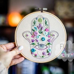 May include: Embroidered hamsa hand with floral details and an evil eye in the center, in a wooden embroidery hoop. The embroidery is done in shades of blue, green, pink, and purple.