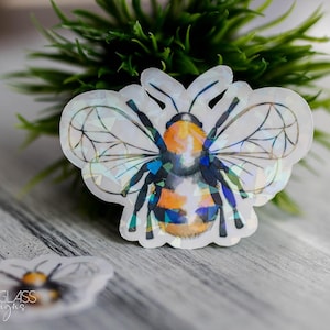 Puede incluir: Una pegatina holográfica de abeja con un diseño detallado, con detalles en naranja, negro y azul. La pegatina está colocada sobre una superficie de madera gris claro con una pequeña planta verde en el fondo.
