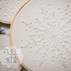 Peut inclure: Gros plan d'un motif de flocon de neige brodé en blanc sur un tissu crème tendu dans un cercle à broder en bois. Le motif présente des points de couture détaillés et un thème hivernal. Les mots "FULL kit" sont visibles.