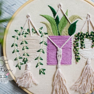 May include: A close-up of a white embroidery hoop with three hanging plants. The plants are embroidered with green leaves and pink flowers. The plants are hanging from a white cord with a tassel at the bottom. The hoop is on a wooden surface.