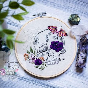 May include: An embroidery hoop featuring a skull design with a moth, spiderweb, and floral accents in purple, orange, and green. The words "FULL kit" are visible. Crystals and greenery are in the background.