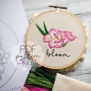 May include: An embroidery hoop with a pink and white flower design and the word "bloom" stitched in black thread. A PDF pattern and embroidery floss are also visible.