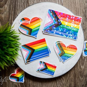 Könnte beinhalten: Eine Sammlung farbenfroher Aufkleber mit Regenbogen- und Pride-Flaggen-Designs. Die Aufkleber haben verschiedene Formen, darunter Herzen und Rechtecke, und sind auf einer runden, hellfarbigen Oberfläche angeordnet. Der Hintergrund ist ein dunkles Holzmaserungsmuster.