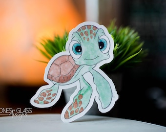 Puede incluir: Una pegatina de acuarela de una tortuga marina de dibujos animados con un cuerpo verde claro, caparazón marrón y grandes ojos azules. La pegatina tiene un borde blanco y el texto "STONES + GLASS designs".