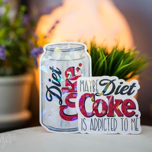 May include: A Diet Coke sticker in the shape of a soda can and a smaller sticker with the text "Maybe Diet Coke is addicted to me." The can sticker has the word "Diet" in blue script.