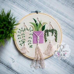 a round embroidery hoop with a floral design, including hanging plants and a pink square. The hoop is placed on a wooden surface, and there is a green plant in the background.