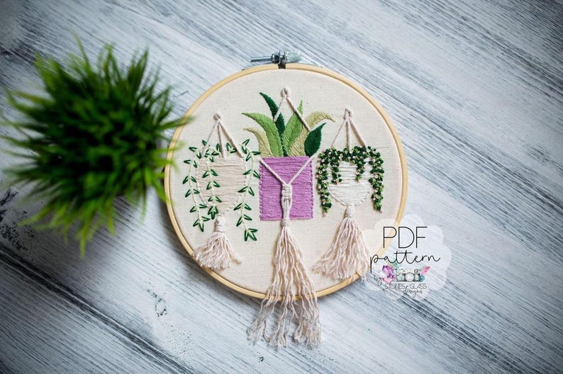 a round embroidery hoop with a floral design, including hanging plants and a pink square. The hoop is placed on a wooden surface, and there is a green plant in the background.