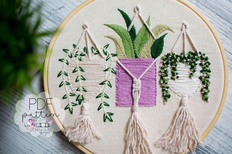 a close-up view of a hand-embroidered piece of art, showcasing three hanging plants with green leaves and white flowers. The embroidery is detailed and intricate, with the plants and flowers appearing to be suspended from a circular hoop. The background is blurred, drawing attention to the embroidered plants and their details.