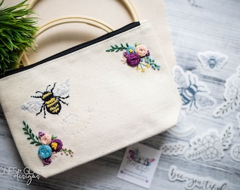 May include: Cream canvas zipped pouch featuring an embroidered bee and floral design. The bee is black and yellow, with pink, purple, and blue flowers. The text "bee-you-tiful" is visible on a sticker.