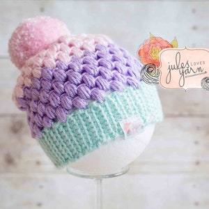 Könnte beinhalten: Eine mehrfarbige gehäkelte Mütze mit einem rosa Bommel. Die Mütze ist in den Farben Rosa, Lila und Grün gehalten. Die Mütze wird auf einem weißen Styropor-Kopf präsentiert. Die Mütze ist mit "jules loves yarn" beschriftet.