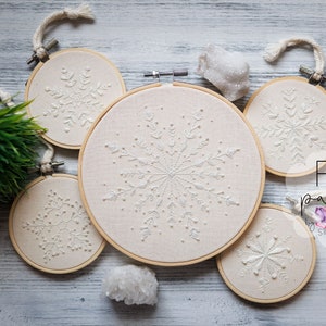 Könnte beinhalten: Fünf Stickrahmen mit weißen Schneeflocken-Designs auf beigefarbenem Stoff. Die Rahmen sind auf einem weißen Holzgrund mit einer kleinen grünen Pflanze und weißen Kristallen angeordnet. Der Text "PDF pattern" ist in der unteren rechten Ecke des Bildes sichtbar.