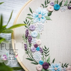 May include: A full embroidery kit featuring a floral wreath design with blue, pink, and purple flowers and green leaves. The kit includes all the materials needed to complete the project, including embroidery floss, needles, and fabric. The kit is labelled "FULL kit by Stones & Glass Designs".
