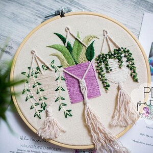 a round embroidery hoop with three hanging plant baskets, each with different types of plants and tassels. The hoop is placed on a wooden surface, and there is a piece of paper with text and a pattern visible in the background.