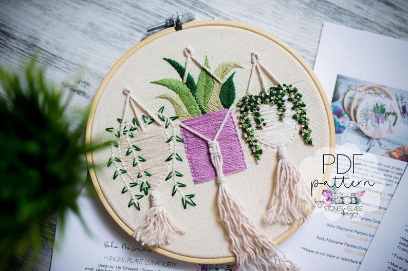 a round embroidery hoop with three hanging plant baskets, each with different types of plants and tassels. The hoop is placed on a wooden surface, and there is a piece of paper with text and a pattern visible in the background.