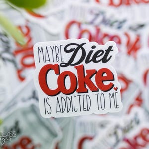 May include: A white sticker with the text "MAYBE Diet Coke IS ADDICTED TO ME" in black and red. The words "Diet Coke" are in a bold red font. The sticker is surrounded by other stickers with the words "Diet Coke" on them.