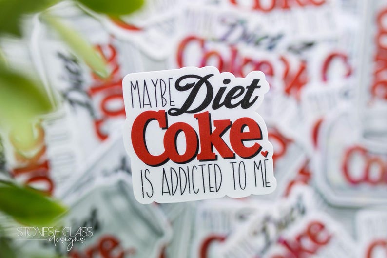 May include: A white sticker with the text "MAYBE Diet Coke IS ADDICTED TO ME" in black and red. The words "Diet Coke" are in a bold red font. The sticker is surrounded by other stickers with the words "Diet Coke" on them.