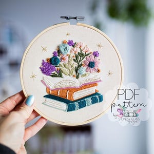 May include: Embroidered hoop art featuring a stack of books with an open book on top, adorned with colorful floral embroidery. The flowers include purple, blue, and pink blooms. The text "PDF pattern" is visible.