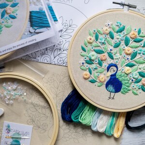 May include: Embroidered peacock design on a beige fabric stretched within a wooden hoop. The peacock is blue, with green and peach floral details. The kit includes embroidery floss and other supplies.