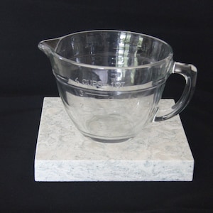 May include: A clear glass measuring cup with a handle, marked with measurements in cups and ounces, sitting on a white square tile.