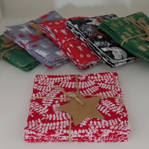 May include: A collection of square fabric coasters in assorted colours and patterns. Designs include red with white leaves, green with gold trees, and grey with white deer. Each stack is tied with twine and a star-shaped tag.