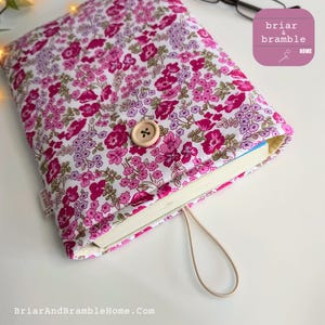 May include: A fabric book sleeve featuring a white background with a dense floral pattern in shades of pink, magenta, and light purple. The sleeve includes a single wooden button closure, an elastic loop, and a small side tag labelled briar & bramble.