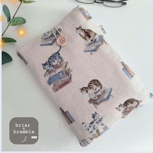 May include: A rectangular beige fabric sleeve featuring a repeating pattern of cats sitting on stacks of books. The sleeve is secured with a light pink button and an elastic loop. A small white tag reads briar & bramble.