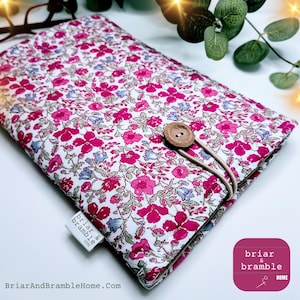 May include: A floral book cover with a button fastening. The cover displays a pattern of pink and blue flowers on a white backdrop. A small label with the text "briar & bramble HOME" is attached. The website BriarAndBrambleHome.com is also visible.