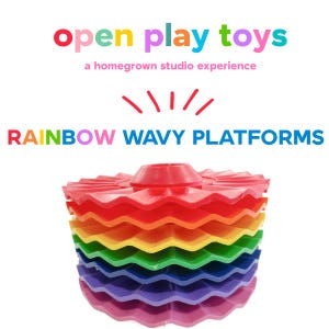 May include: A stack of circular, wavy-edged platforms in rainbow colors including red, orange, yellow, green, blue, purple, and pink. The text above reads open play toys a homegrown studio experience and RAINBOW WAVY PLATFORMS.