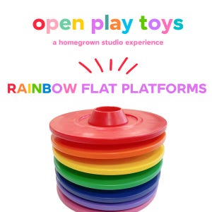 Può includere: Una pila di piattaforme piatte circolari arcobaleno in rosso, arancione, giallo, verde, blu, indaco e violetto. Il testo sopra recita open play toys, a homegrown studio experience e RAINBOW FLAT PLATFORMS con un arco grafico rosso.