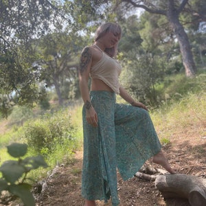 May include: A woman wearing a white crop top and patterned pants stands in a forest setting. She is barefoot and has a tattoo on her arm.