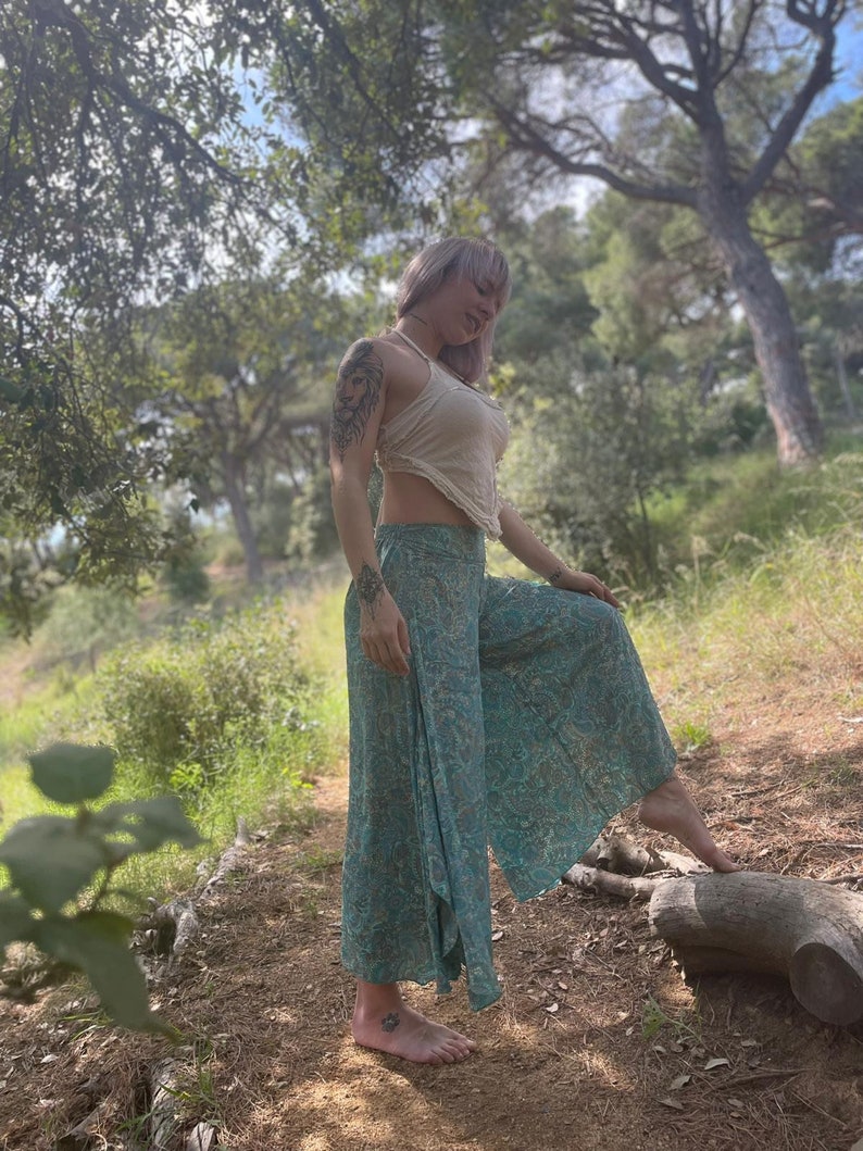 May include: A woman wearing a white crop top and patterned pants stands in a forest setting. She is barefoot and has a tattoo on her arm.