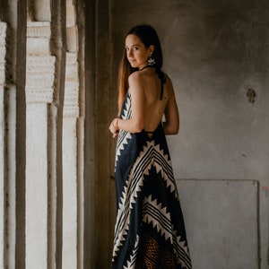 May include: A woman wearing a black and white patterned maxi dress with a halter neckline. She is standing in a doorway with white pillars.