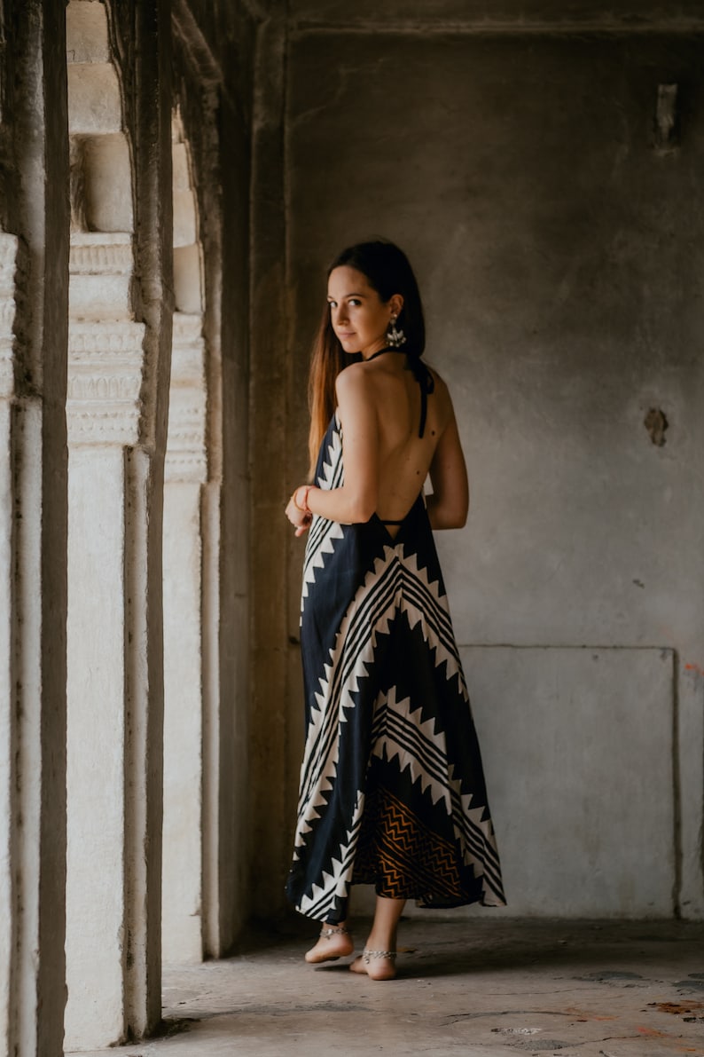 May include: A woman wearing a black and white patterned maxi dress with a halter neckline. She is standing in a doorway with white pillars.