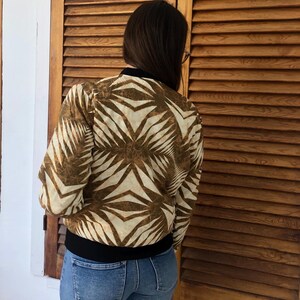 Puede incluir: Una chaqueta bomber con estampado marr&oacute;n y blanco con una banda negra. La chaqueta est&aacute; hecha de una tela suave y sedosa.