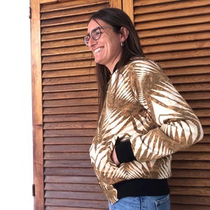 Puede incluir: Una mujer lleva una chaqueta bomber con estampado marr&oacute;n y blanco con ribetes negros. Est&aacute; sonriendo y mirando hacia un lado. La chaqueta tiene una cremallera y un cuello de pie.