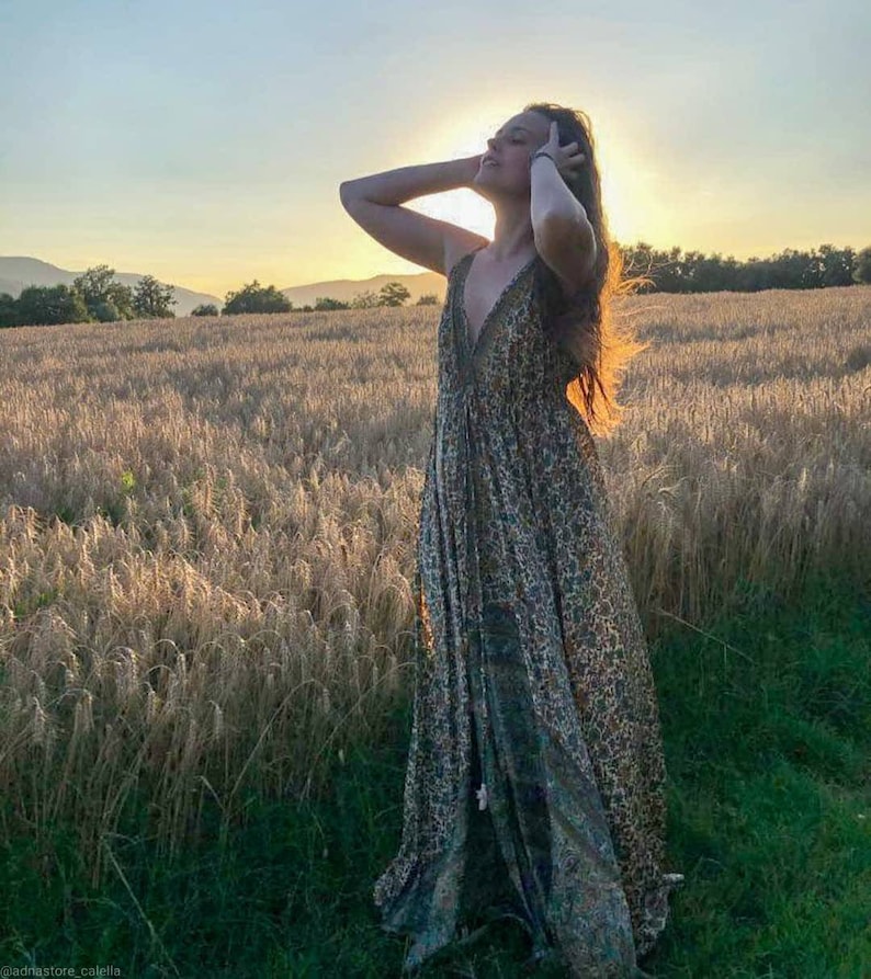 May include: A woman wearing a long, flowing, patterned maxi dress stands in a field of tall, golden wheat. The sun is setting behind her, creating a warm glow.