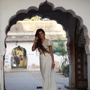 Peut inclure: Une femme aux longs cheveux bruns porte une jupe blanche enroul&eacute;e et un haut blanc avec des d&eacute;tails en dentelle. Elle se tient dans une porte avec une ouverture en arc. La porte est en pierre blanche et a un design indien traditionnel.