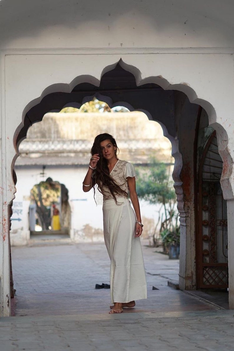 Peut inclure: Une femme aux longs cheveux bruns porte une jupe blanche enroul&eacute;e et un haut blanc avec des d&eacute;tails en dentelle. Elle se tient dans une porte avec une ouverture en arc. La porte est en pierre blanche et a un design indien traditionnel.