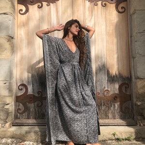 May include: A woman wearing a long, flowing black and white patterned kaftan dress with a deep V-neckline. She is standing in front of a wooden door with ornate iron hinges.