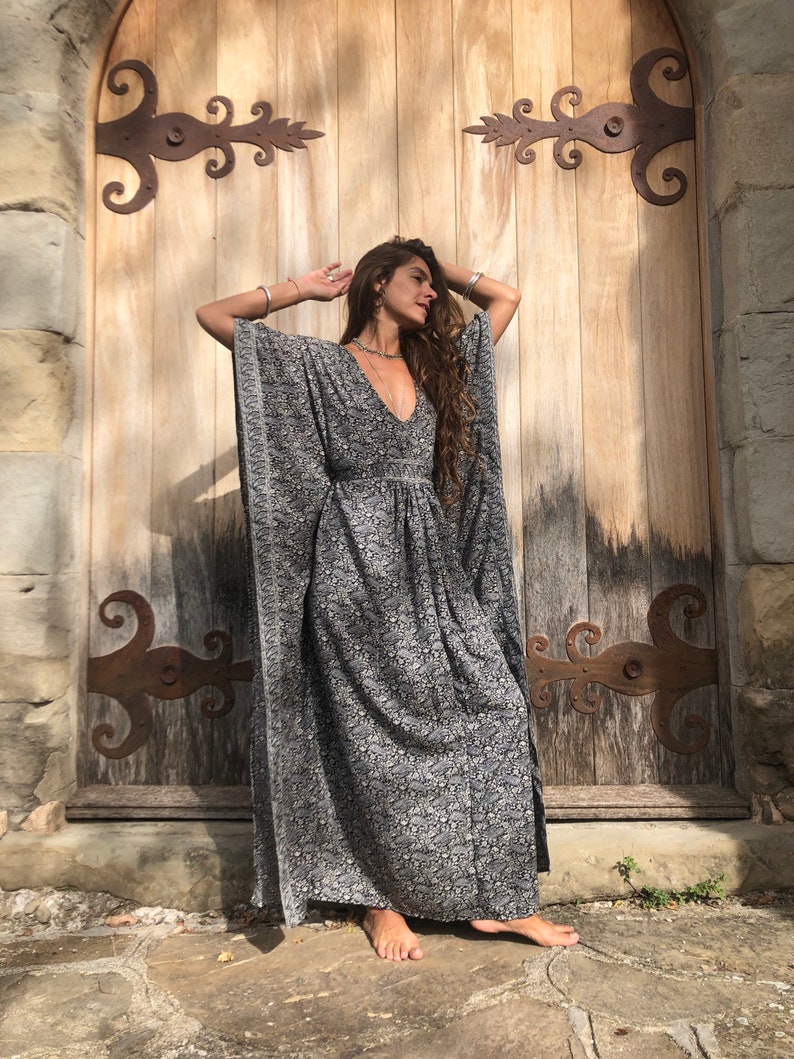 May include: A woman wearing a long, flowing black and white patterned kaftan dress with a deep V-neckline. She is standing in front of a wooden door with ornate iron hinges.