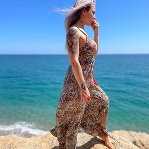 May include: A woman with purple hair wearing a long, flowing floral dress with a lion tattoo on her arm. She is standing on a rocky beach with the ocean in the background.