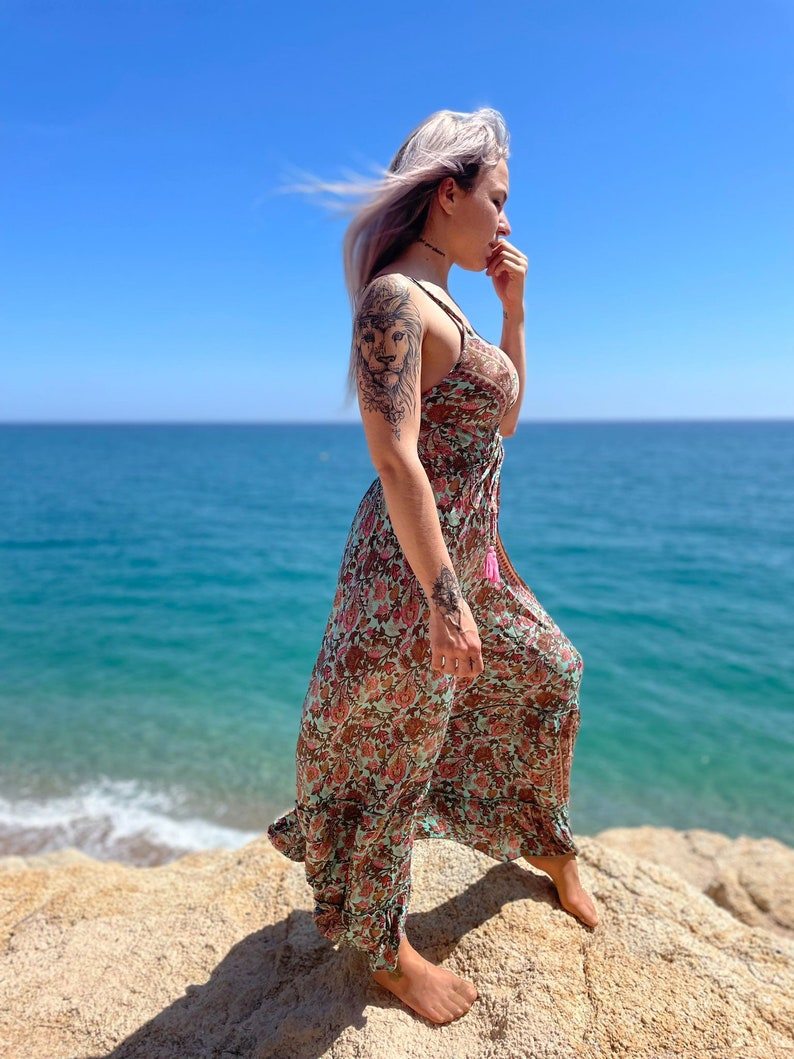 May include: A woman with purple hair wearing a long, flowing floral dress with a lion tattoo on her arm. She is standing on a rocky beach with the ocean in the background.